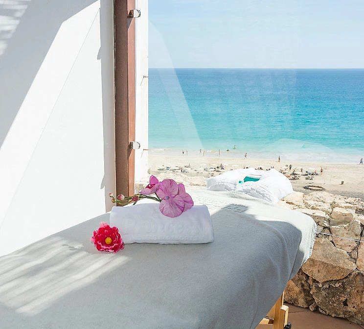 Cama junto a la ventana con flores y vista al mar desde el balc&oacute;n
