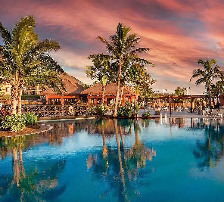 Piscina azul con palmeras y casas al atardecer, cielo rosado y reflejos