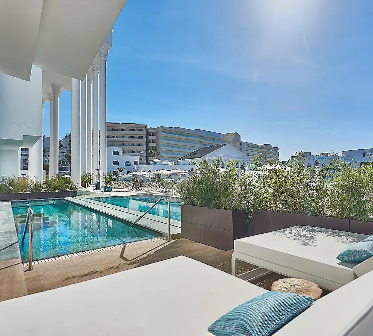 Piscina y terraza con tumbonas blancas, vista a edificios bajo cielo azul