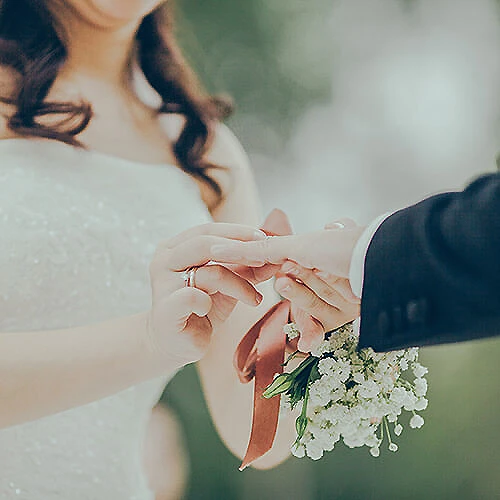 Pareja intercambiando anillos de boda, con ramo de flores al fondo.