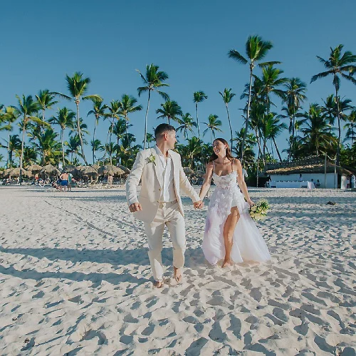 Pareja con vestido de novia y novio caminando en playa con palmeras