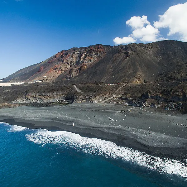 Playa negra con olas y acantilados volc&aacute;nicos bajo un cielo azul.