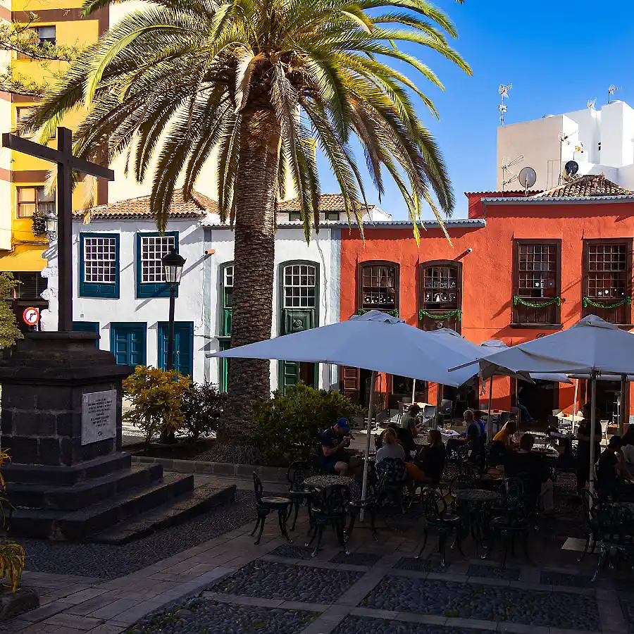 Plaza con palmera, edificios coloridos y mesas bajo sombrillas, p&uacute;blico reunido