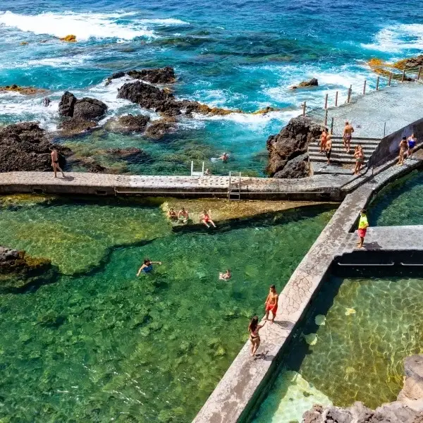 Personas caminando y nadando en una piscina natural junto al mar y rocas.