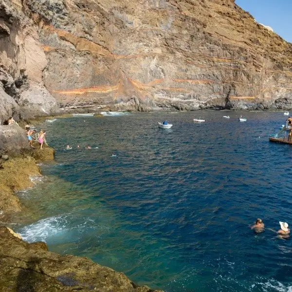 Bah&iacute;a azul con botes y personas nadando junto a acantilados rocosos.