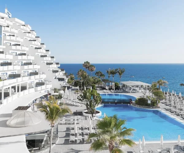 Hotel junto al mar con piscina azul, palmeras y tumbonas blancas