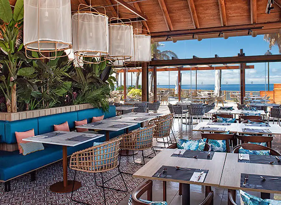 Terraza de restaurante con mesas, sillas y l&aacute;mparas colgantes frente al mar
