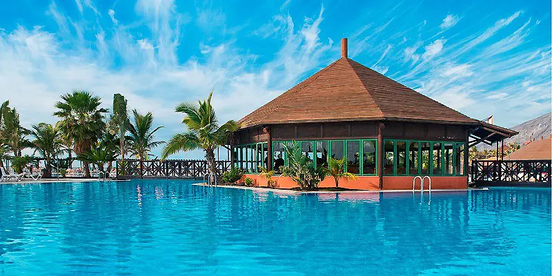 Piscina azul frente a un pabell&oacute;n tropical con palmeras y cielo despejado