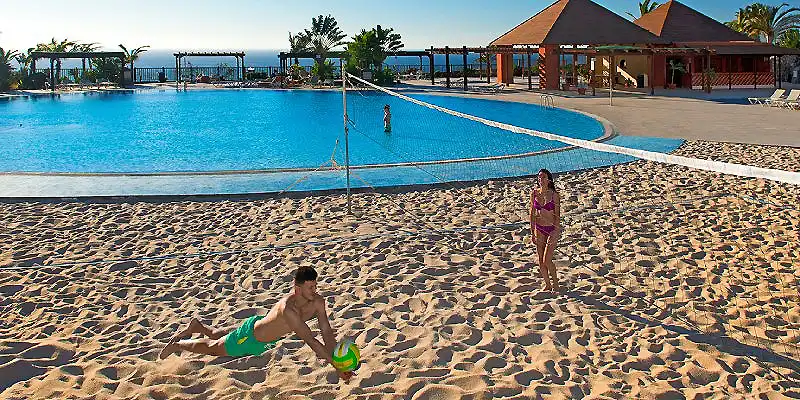 Playa con piscina, ni&ntilde;o jugando con pelota y caba&ntilde;as al fondo.