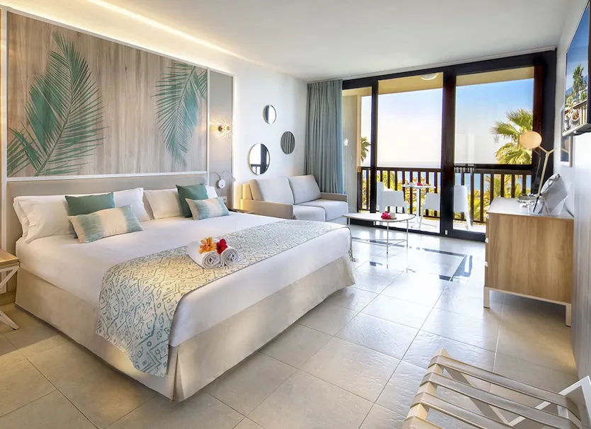 Dormitorio moderno con cama, balc&oacute;n y vista a palmeras bajo luz natural