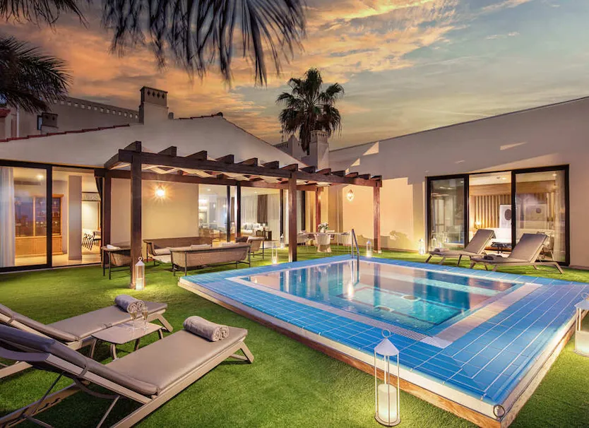 Piscina con tumbonas en jard&iacute;n, palmeras y atardecer junto a una casa.