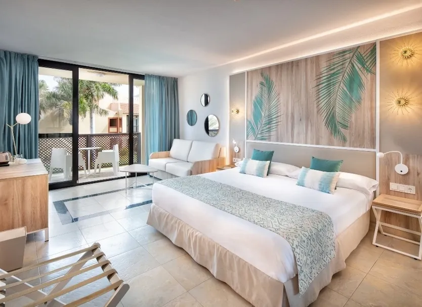 Dormitorio moderno con cama blanca, sof&aacute;, palmeras y grandes ventanales.