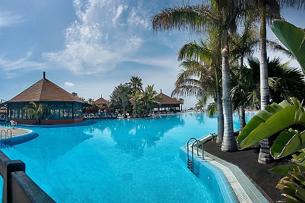 Piscina tropical con palmeras y caba&ntilde;as al fondo bajo un cielo azul