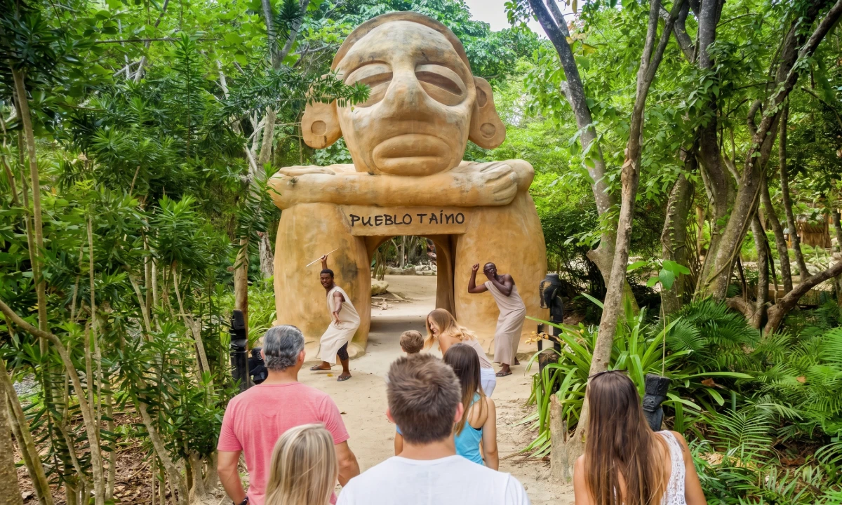 Touristas caminan frente a una estatua de piedra ta&iacute;na en un jard&iacute;n tropical.