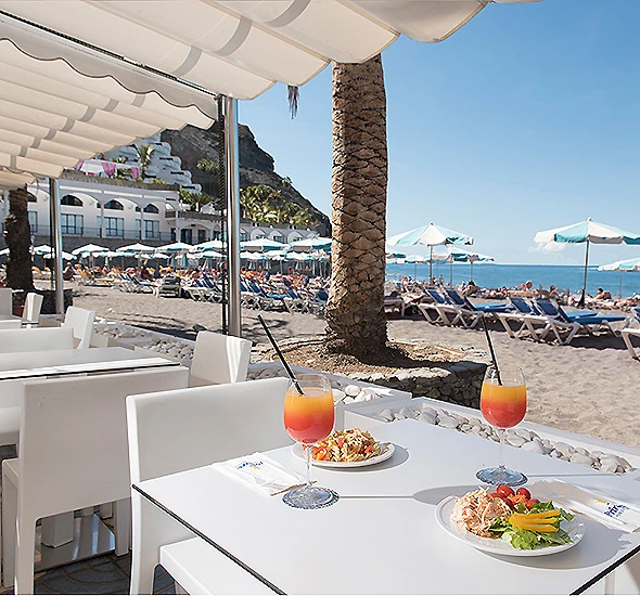 Terraza frente al mar con palmeras, mesas y c&oacute;cteles en la playa.