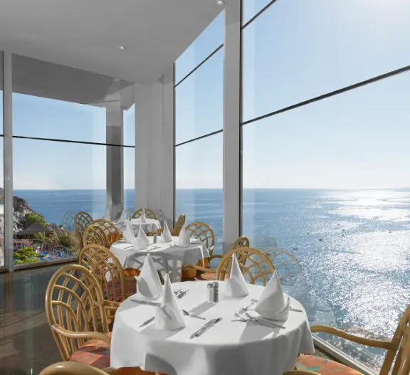 Mesa con sillas junto a ventanales con vista al mar y el paisaje costero.