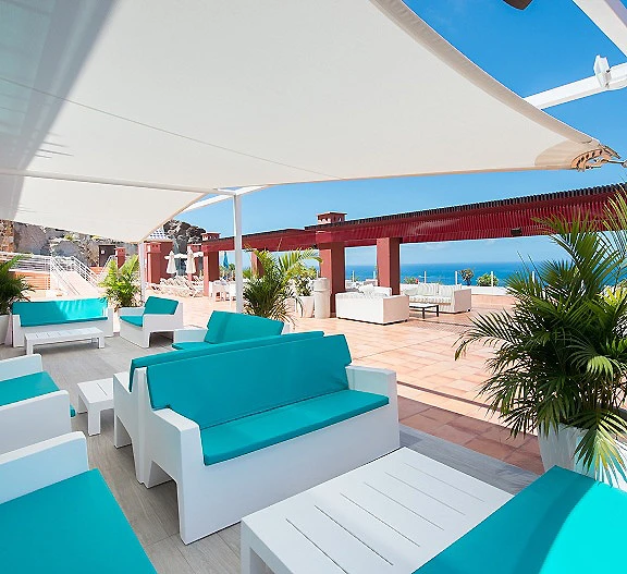 Terraza con sof&aacute;s azules bajo toldos y vistas al mar desde el hotel.