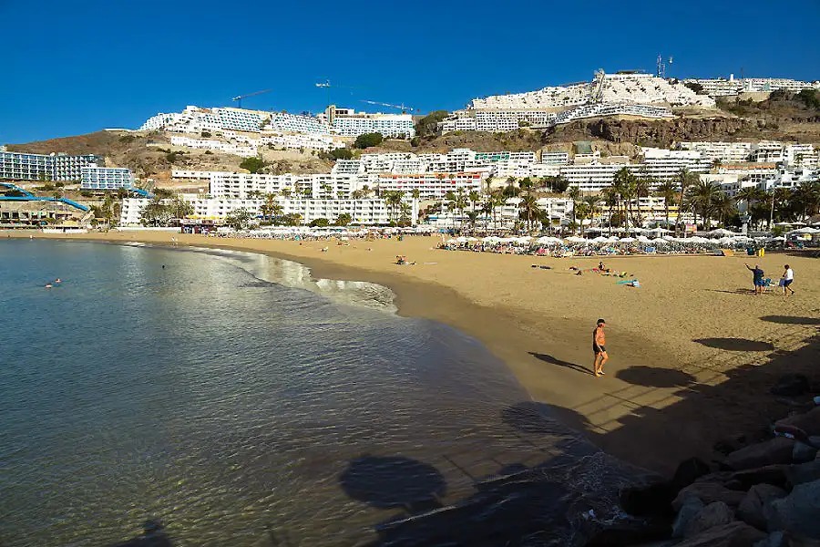 Playa soleada con arena, olas y edificios en acantilado al fondo.
