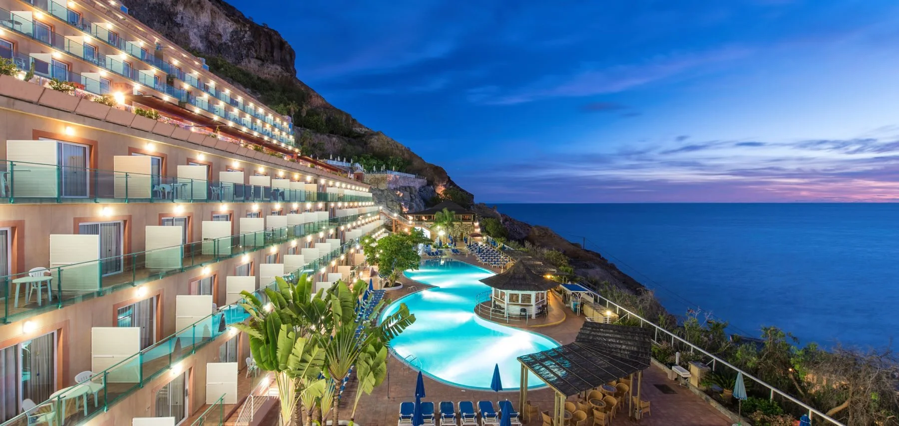 Hotel junto al mar con piscina iluminada y balcones en la ladera, al atardecer