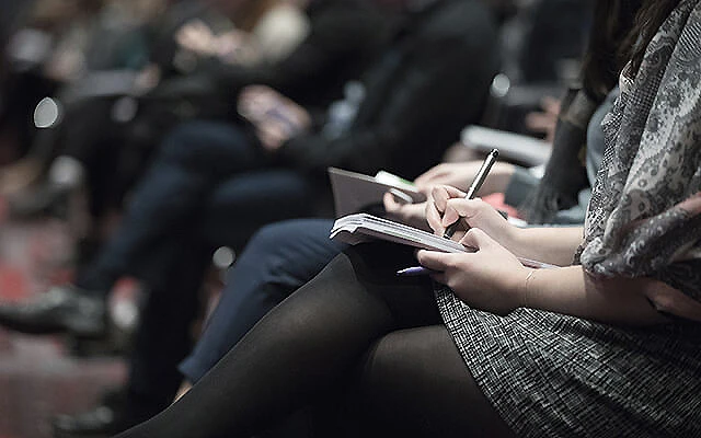 Persona escribiendo en un cuaderno en un evento, con p&uacute;blico al fondo