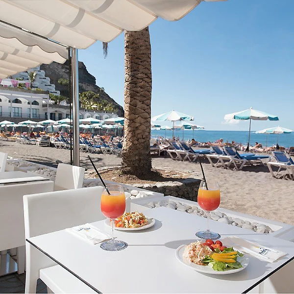 Desayuno y c&oacute;cteles en una terraza frente a la playa, con sombrillas y hamacas.