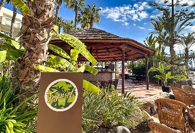 Jard&iacute;n tropical con palmeras, caba&ntilde;a y letrero verde junto a plantas