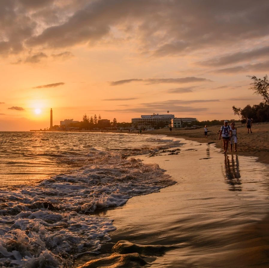 Atardecer en la playa con olas, faro y personas caminando por la orilla.