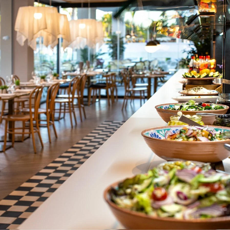 Buffet con ensaladas en cuencos, comedor con mesas y sillas al fondo.