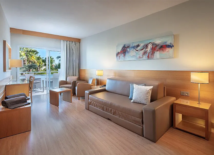 Sala de estar moderna con sof&aacute;, l&aacute;mparas y balc&oacute;n con palmeras al fondo.