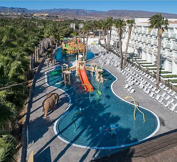 Piscina y tobog&aacute;n en hotel, palmeras y patio con sillas y monta&ntilde;as al fondo.