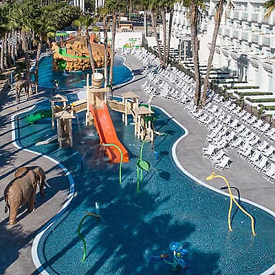 Piscina infantil con tobog&aacute;n y parque acu&aacute;tico en un hotel con palmeras.