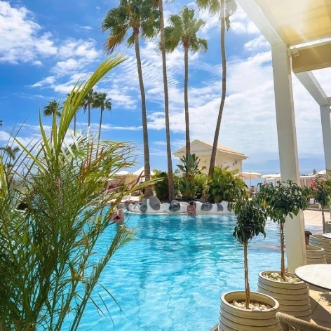 Piscina azul con palmeras y cielo despejado junto a una terraza.