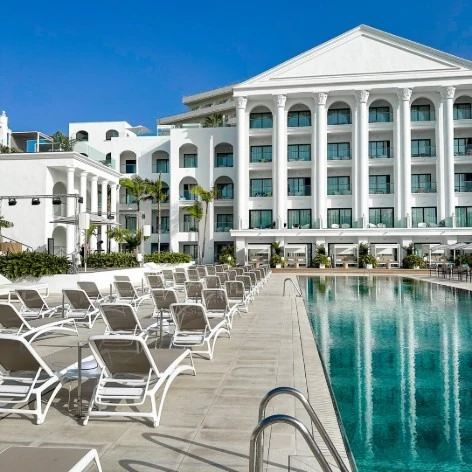Piscina junto a edificio blanco con columnas y tumbonas bajo cielo azul