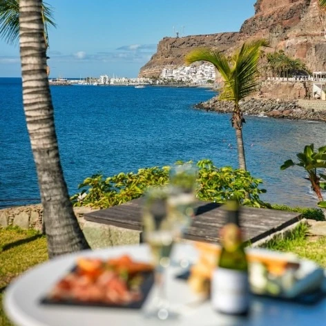 Vista del mar y costa con palmeras y mesas de jard&iacute;n en primer plano.