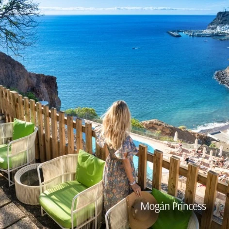 Mujer frente al mar en terraza con sillas verdes, vista a acantilados en Mog&aacute;n