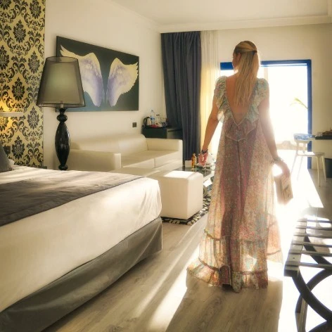 Mujer de pie junto a una ventana, en una habitaci&oacute;n de hotel con cama.