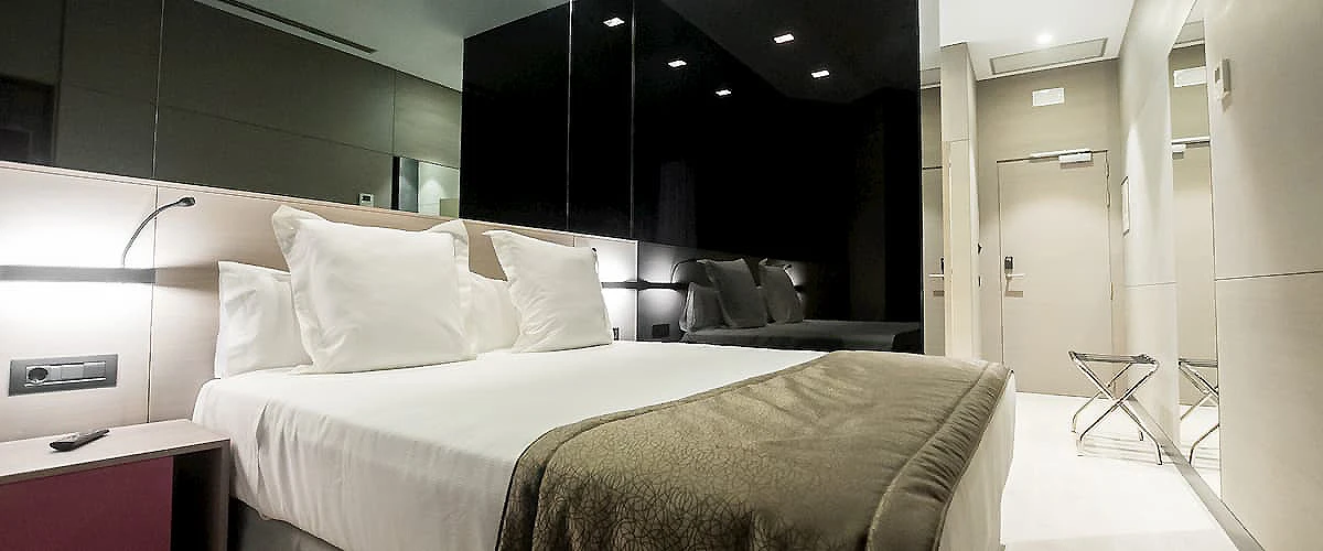 Habitaci&oacute;n de hotel moderna con cama doble, cortinas y sillones grises.