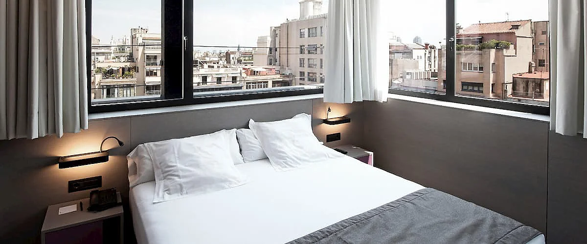 Dormitorio moderno con cama blanca y ventanas amplias con vista urbana, luz natural.