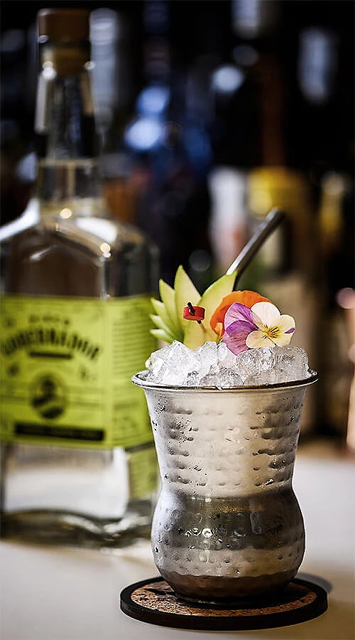 C&oacute;ctel con hielo en vaso met&aacute;lico, flor y pajita, fondo borroso de bar