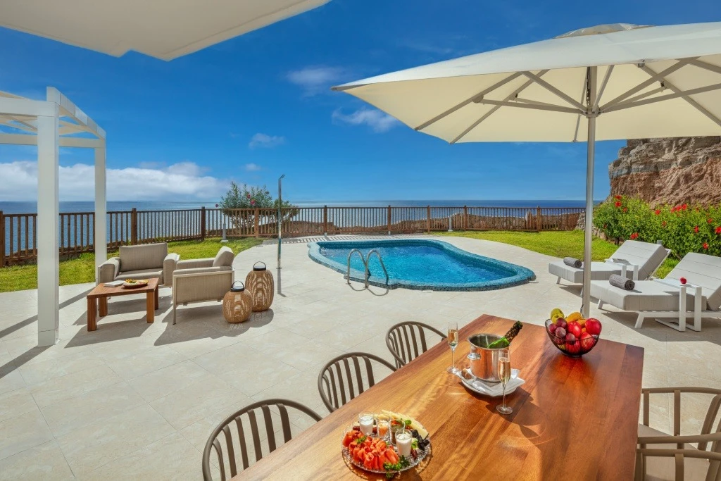 Terraza con piscina azul, muebles y mesa con frutas, vista al mar.