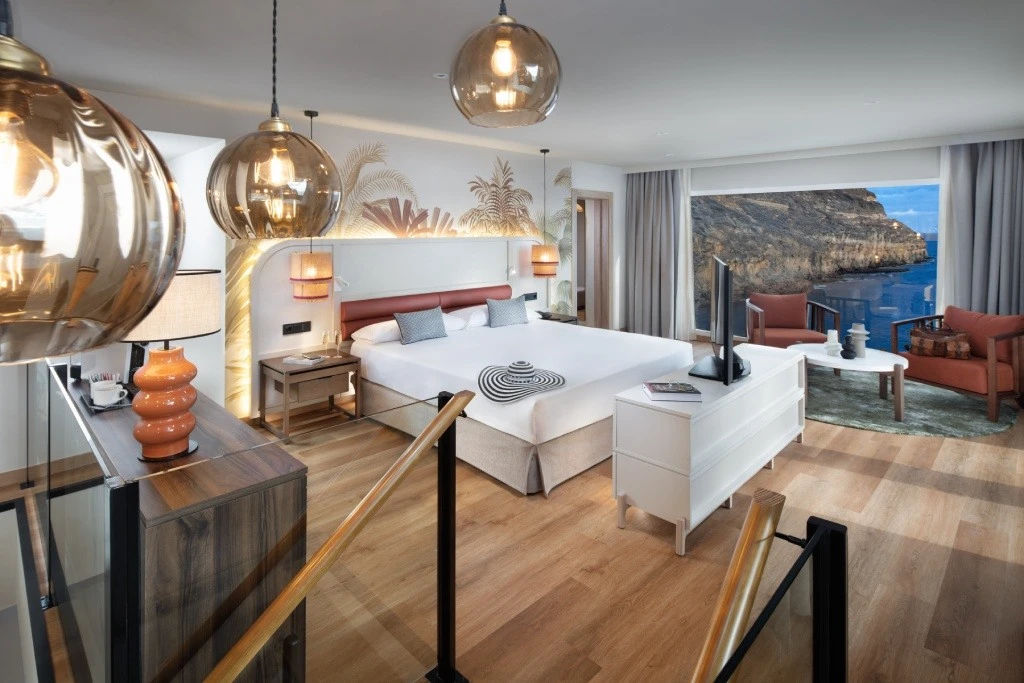 Suite hotelera moderna con cama, l&aacute;mparas colgantes y vista al mar.