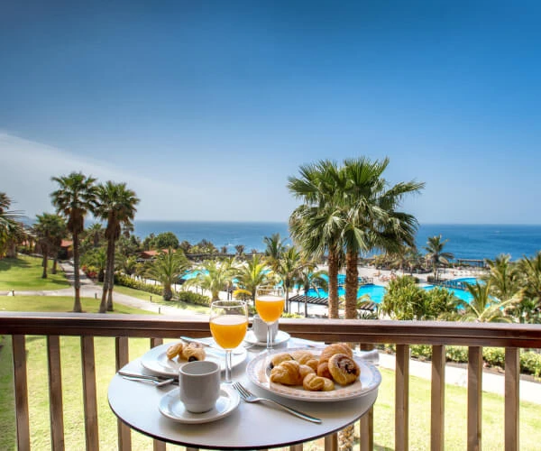 Desayuno en balc&oacute;n con croissants y jugo frente al mar y palmeras