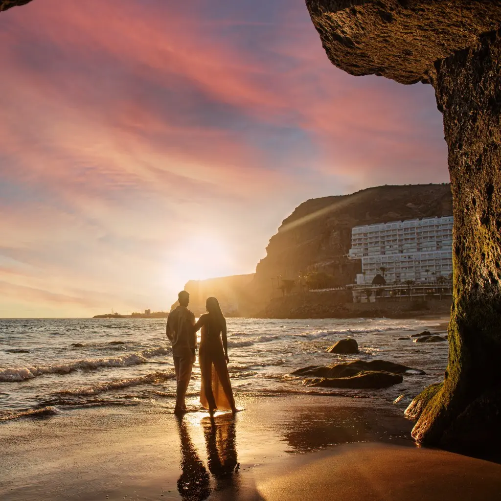 Pareja camina en la playa al atardecer junto a un acantilado y hotel.