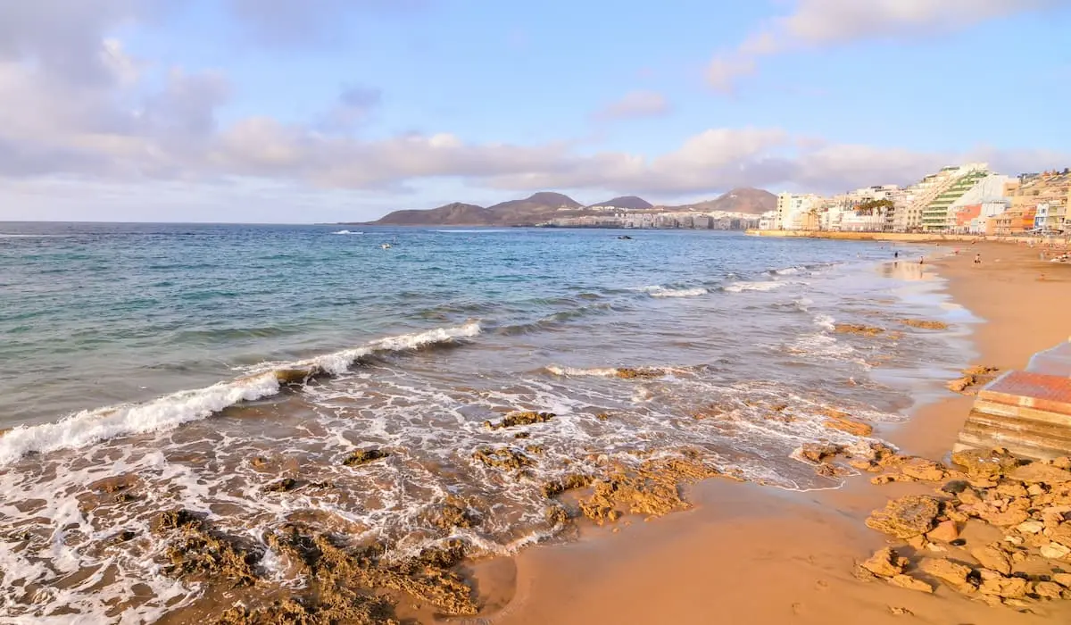 Playa del Atl&aacute;ntico con oleaje y edificios costeros al fondo, cielo nublado