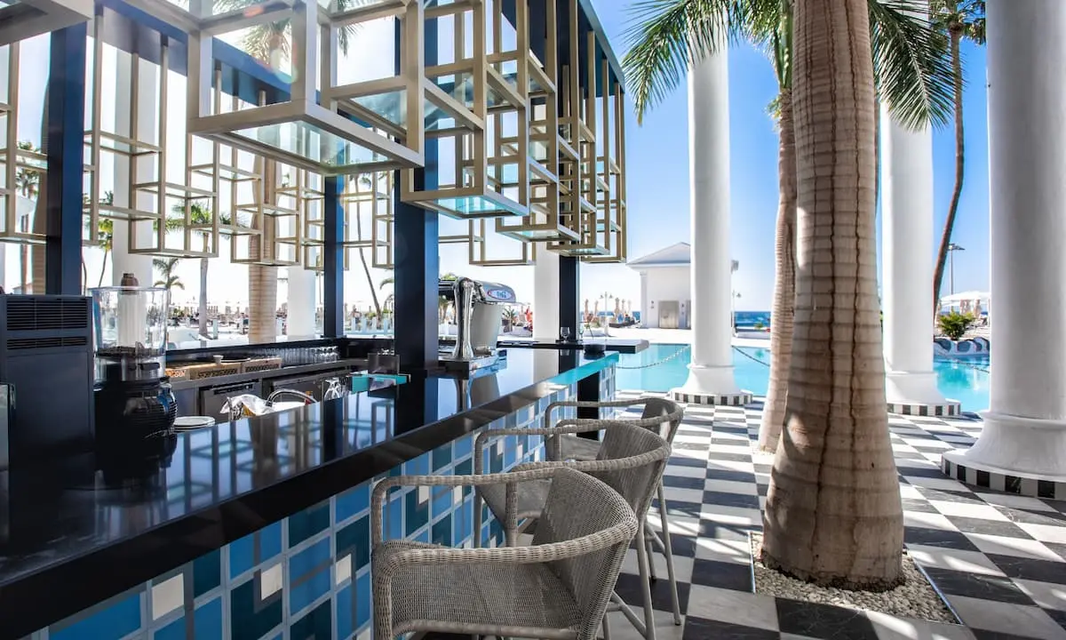 Terraza junto a piscina con columnas blancas, palmeras y mesa de servicio azul