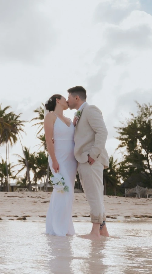 Pareja de reci&eacute;n casados bes&aacute;ndose en la playa, bajo el cielo nublado
