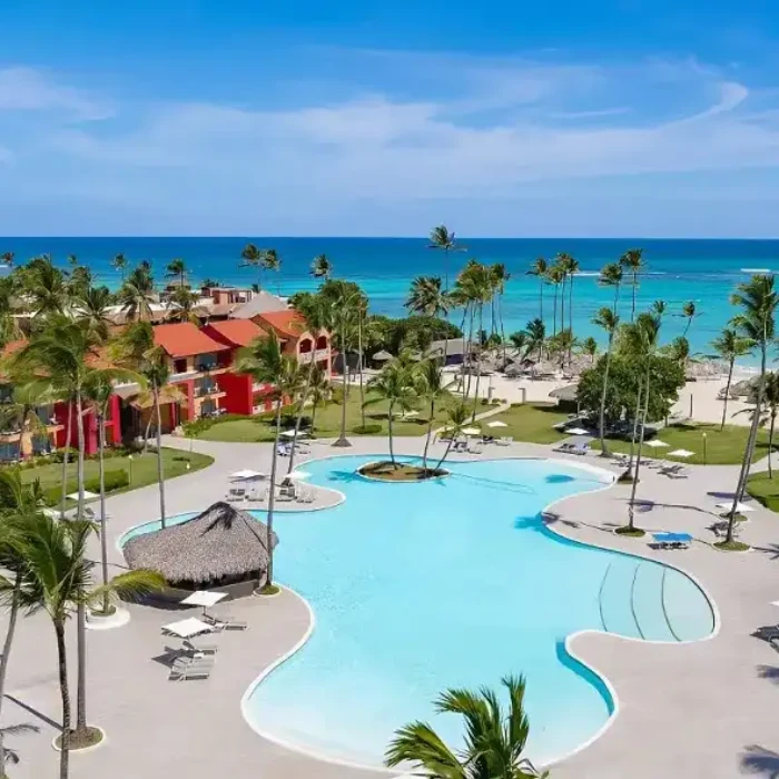 Complejo tur&iacute;stico con palmeras, piscina y vista al mar azul.
