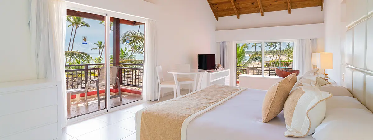 Habitaci&oacute;n de hotel luminosa con cama, balc&oacute;n y vista al mar y palmeras