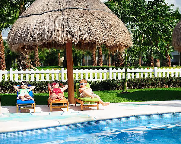 Tres ni&ntilde;os en camastros junto a una piscina con sombrillas de paja.