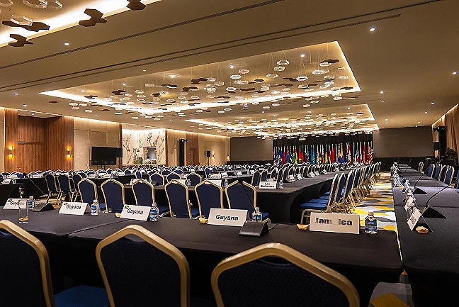 Sala de conferencias con mesas, sillas y banderas en el escenario.